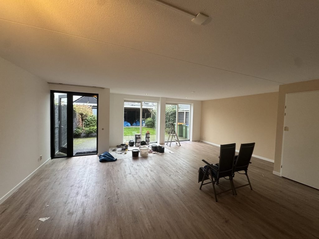 Ruime woonkamer met tuindeuren en een houten vloer, klaar om te behangen, met verfemmers op de vloer